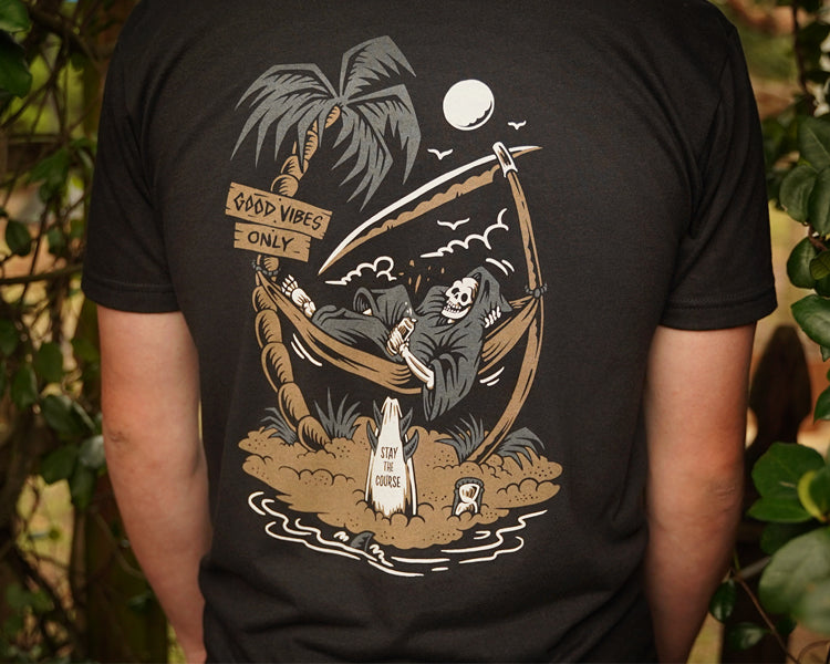 Person wearing a black t-shirt with a graphic design of a skeleton in a hammock, palm trees, and a moon.