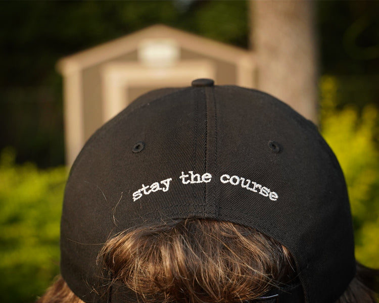 Black cap with 'stay the course' text worn by a person, with a blurred background of greenery and a building.