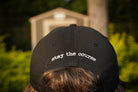 Black cap with 'stay the course' text worn by a person, with a blurred background of greenery and a building.