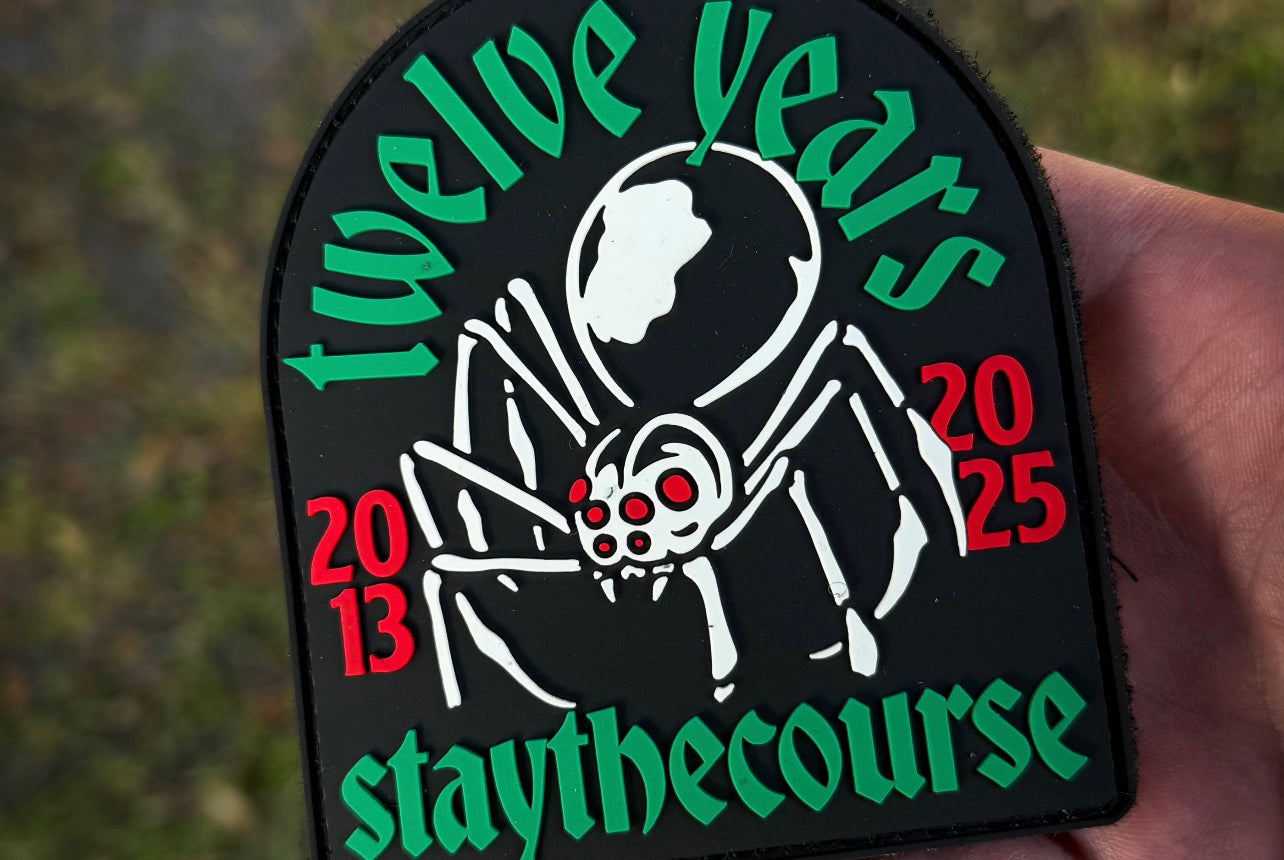 Patch with 'twelve years 2013-2025 staythecourse' text held by a hand against a blurred green background