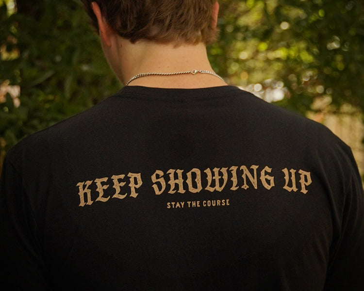 A person viewed from behind wearing a black t-shirt with the phrase 'KEEP SHOWING UP' and 'STAY THE COURSE' printed in a tan serif font, set against a blurred outdoor background of green foliage.
