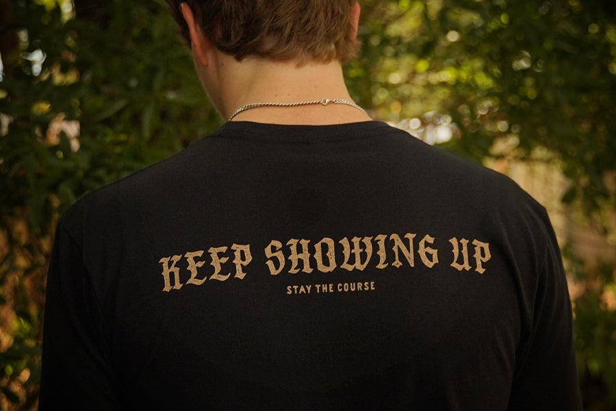 A person viewed from behind wearing a black t-shirt with the phrase 'KEEP SHOWING UP' and 'STAY THE COURSE' printed in a tan serif font, set against a blurred outdoor background of green foliage.