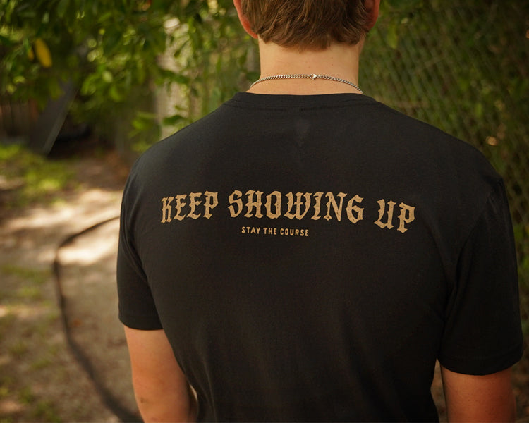 A person viewed from behind wearing a black t-shirt with 'KEEP SHOWING UP' and 'STAY THE COURSE' printed in gold on the back, set against a blurred outdoor background.