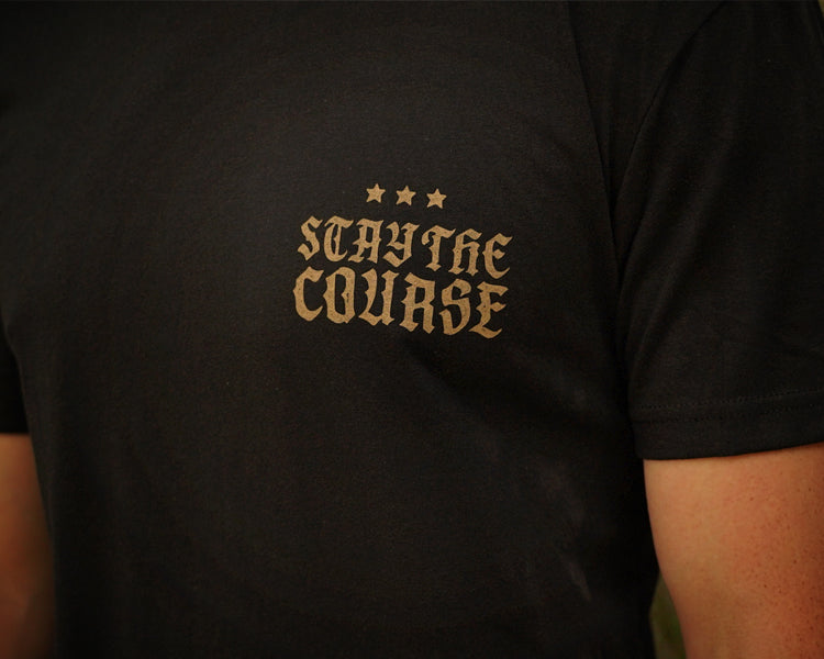 A close-up of a person wearing a black t-shirt with a gold printed graphic on the chest that reads 'STAY THE COURSE' with three stars above it.