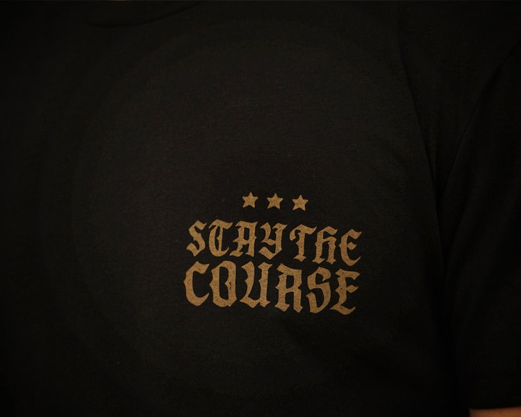 Close-up of a black t-shirt featuring the phrase 'STAY THE COURSE' in a gold gothic font, accented by three gold stars above, set against a dark background.