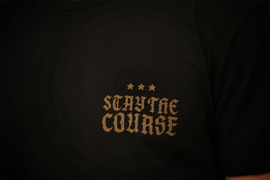 Close-up of a black t-shirt featuring the phrase 'STAY THE COURSE' in a gold gothic font, accented by three gold stars above, set against a dark background.