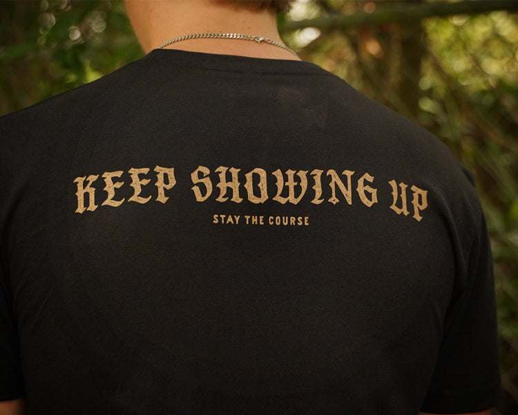 A person viewed from behind wearing a black T-shirt with the phrase 'KEEP SHOWING UP' and smaller text 'STAY THE COURSE' printed in tan across the back, set against a blurred outdoor background.