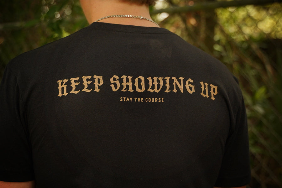 A person viewed from behind wearing a black T-shirt with the phrase 'KEEP SHOWING UP' and smaller text 'STAY THE COURSE' printed in tan across the back, set against a blurred outdoor background.