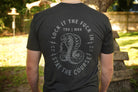 Person wearing a black t-shirt with a snake graphic and text on the back, standing outdoors.