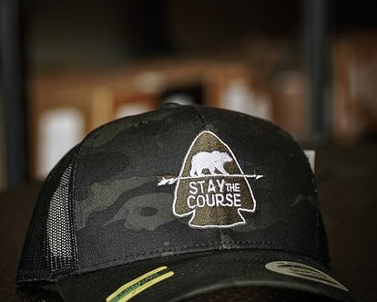 Black multicam trucker hat with 'Stay the Course' embroidered bear graphic on a blurred background