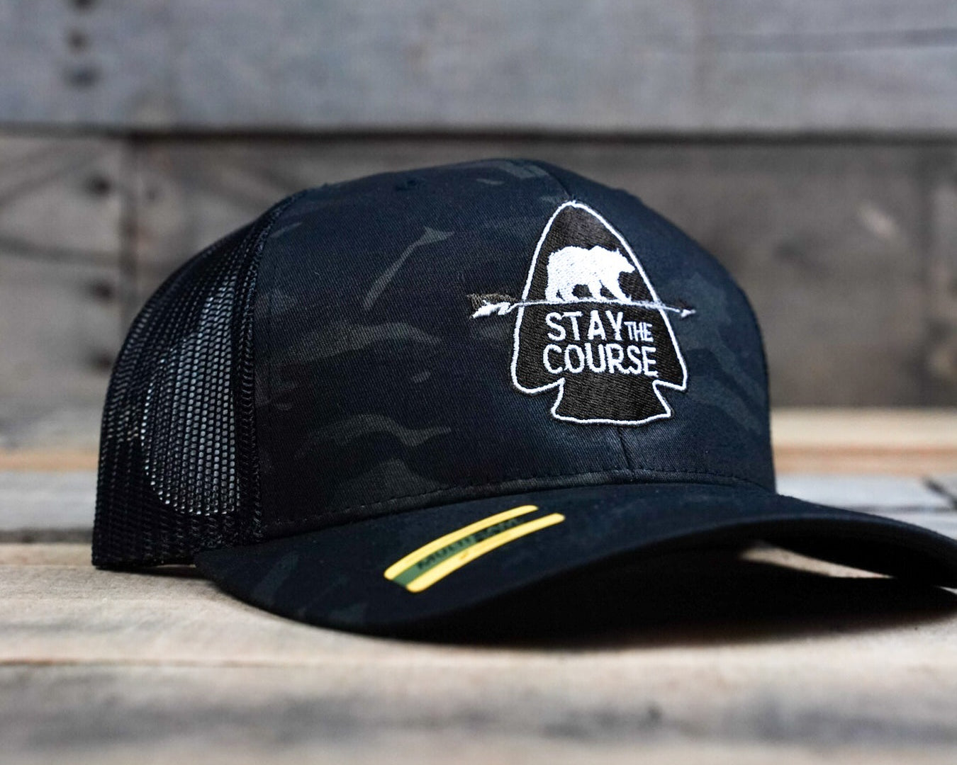 Black multicam cap with 'Stay the Course' logo on a wooden surface