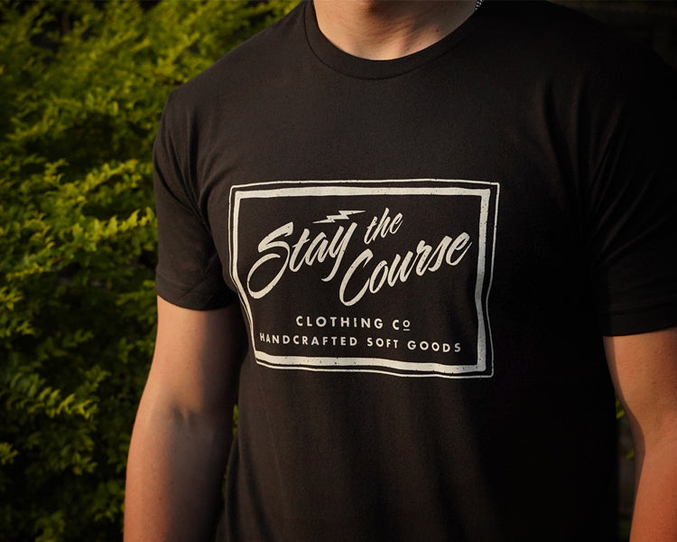 Person wearing a black t-shirt with 'Stay the Course' logo against a blurred green background