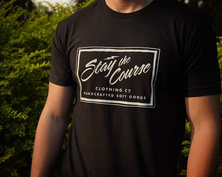 Person wearing a black t-shirt with a logo and text in an outdoor setting