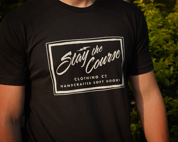 Person wearing a black t-shirt with 'Stay the Course' branding against a blurred green background