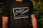 Person wearing a black t-shirt with 'Stay the Course' branding against a blurred green background
