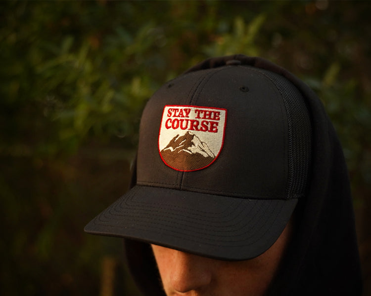 A person wearing a hat with a shield-shaped embroidered patch that reads 'STAY THE COURSE' above a mountain graphic, photographed outdoors with blurred green foliage in the background.