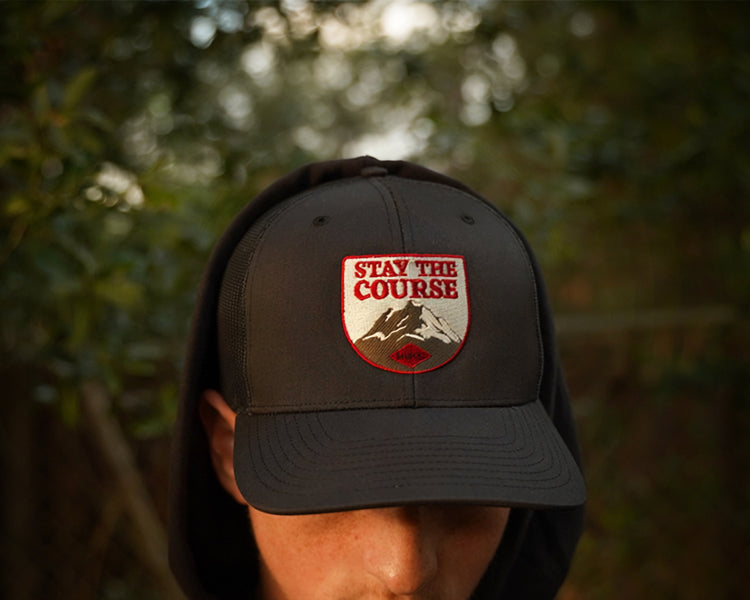 A person wearing a hat with a shield-shaped embroidered patch that reads 'STAY THE COURSE' above a mountain graphic, photographed outdoors with blurred green foliage in the background.