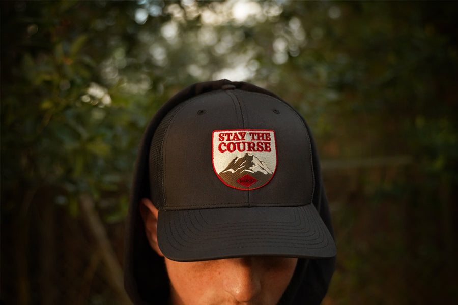A person wearing a hat with a shield-shaped embroidered patch that reads 'STAY THE COURSE' above a mountain graphic, photographed outdoors with blurred green foliage in the background.