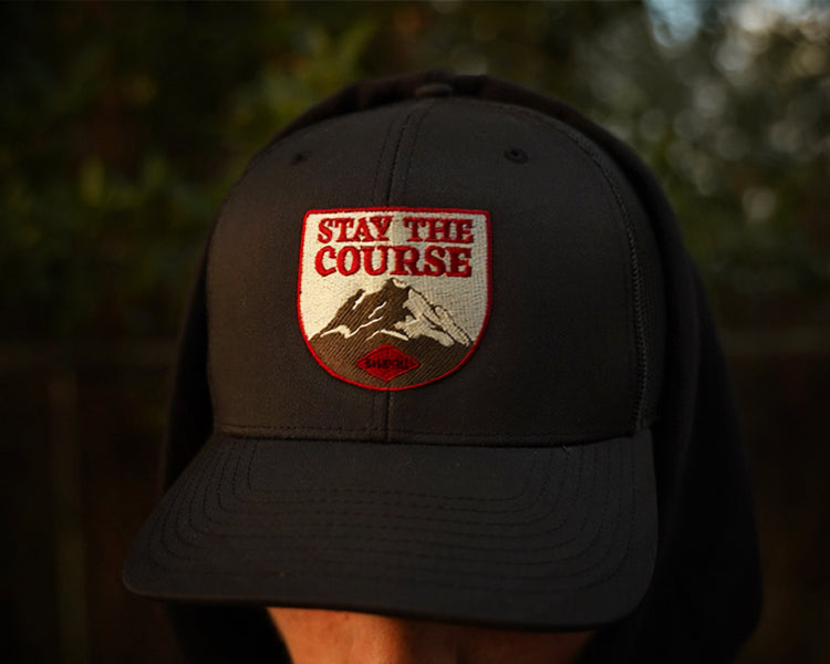 Person wearing a black trucker hat with patch style embroidery of a mountain with stay the course