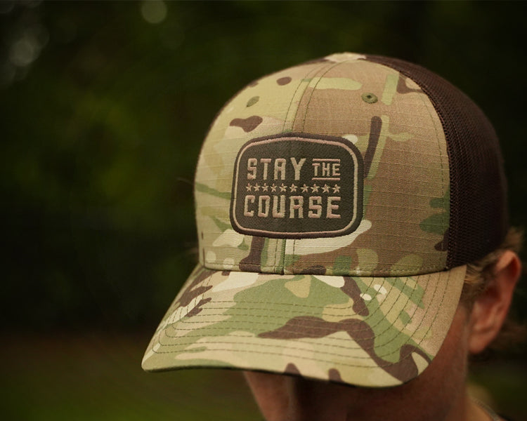 Camouflage cap with 'Stay the Course' patch worn by a person outdoors