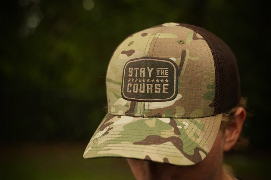 Camouflage cap with 'Stay the Course' patch worn by a person outdoors