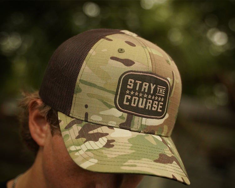 Camouflage cap with 'Stay the Course' logo worn by a person outdoors