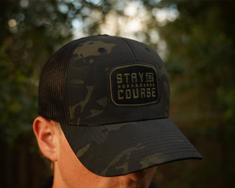 Person wearing a multicam black tactical trucker hat with "Stay the Course" embroidered in black and green, in an outdoor setting