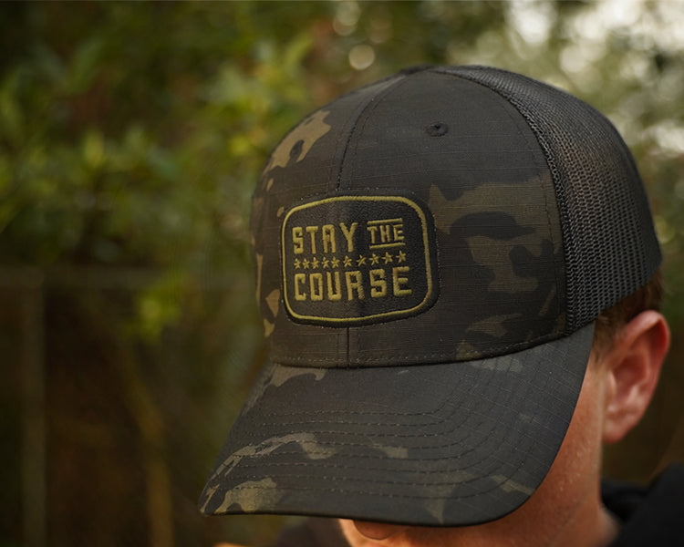 Person wearing a multicam black tactical trucker hat with "Stay the Course" embroidered in black and green, in an outdoor setting