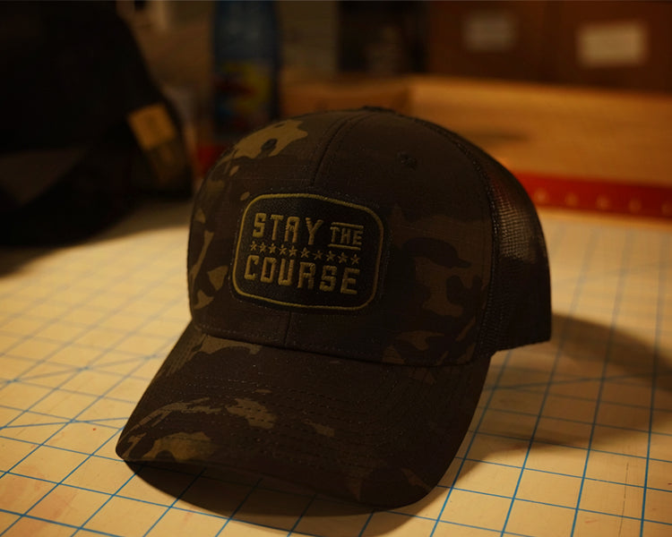 a multicam black tactical trucker hat with "Stay the Course" embroidered in black and green, on a cutting mat