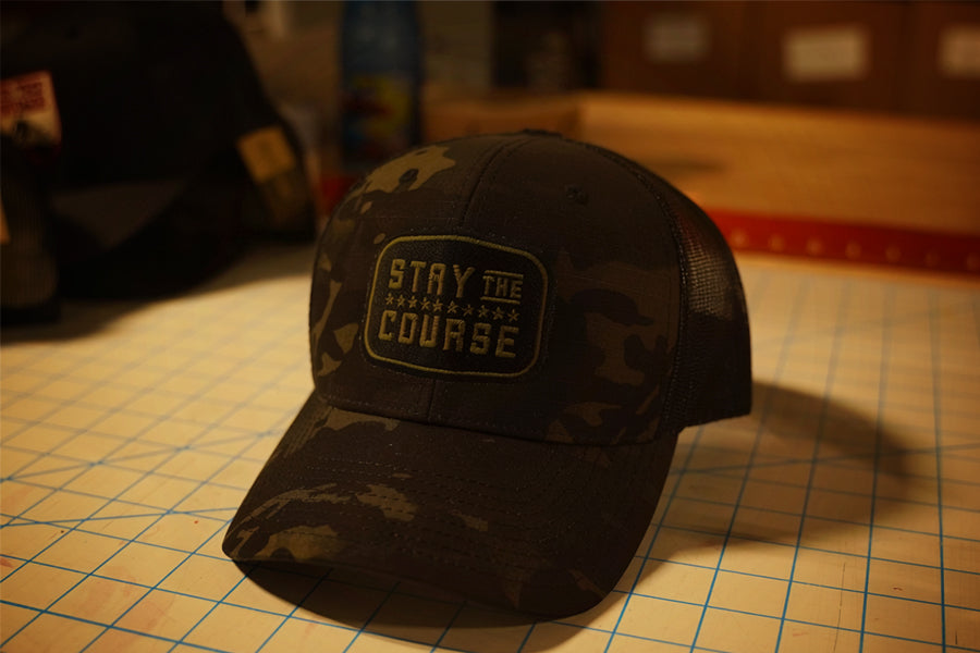 a multicam black tactical trucker hat with "Stay the Course" embroidered in black and green, on a cutting mat