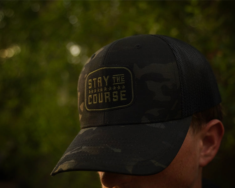 Person wearing a multicam black tactical trucker hat with "Stay the Course" embroidered in black and green, in an outdoor setting