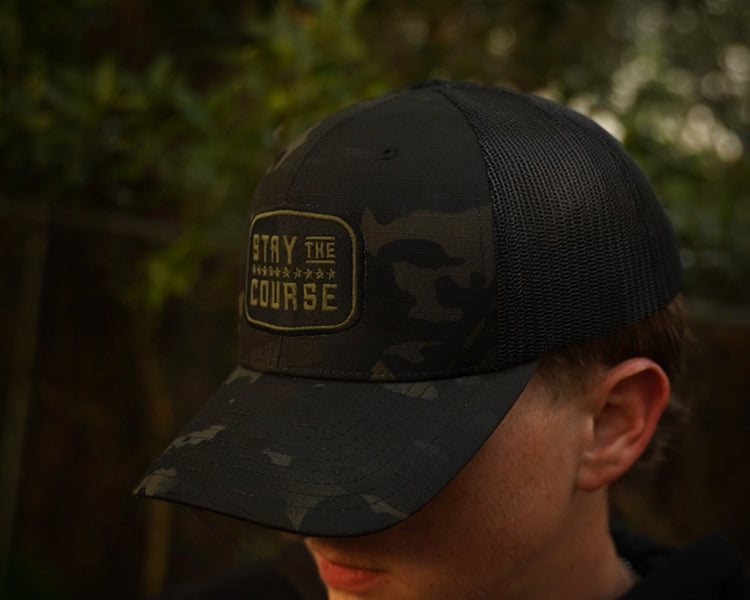Person wearing a multicam black tactical trucker hat with "Stay the Course" embroidered in black and green, in an outdoor setting