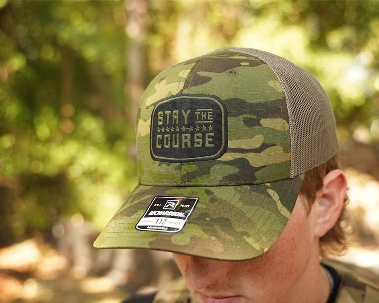 Camouflage cap with 'Stay the Course' patch worn by a person in a forest setting