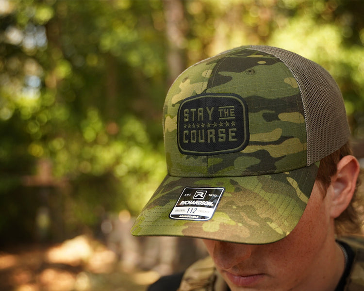 Camouflage cap with 'Stay the Course' patch worn by a person in a forest setting