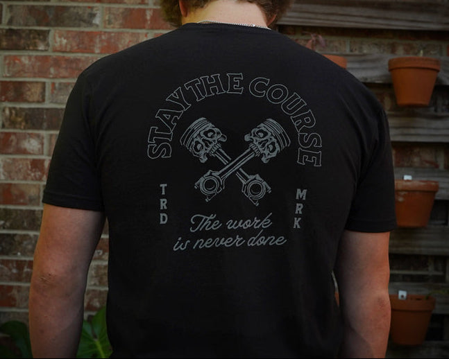 Person wearing a black t-shirt with a graphic design and text, standing in front of a brick wall.