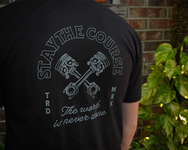 Black t-shirt with 'Stay the Course' graphic and text on the back, worn by a person standing against a brick wall.