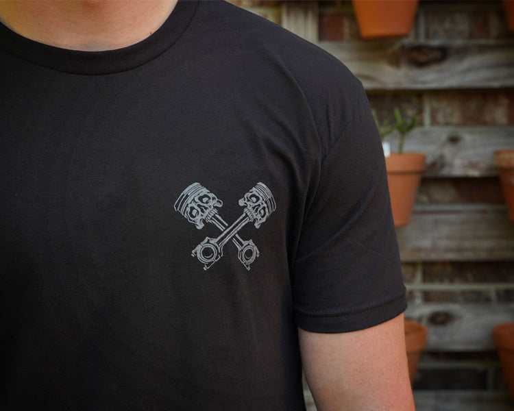 Person wearing a black t-shirt with a gray piston design, standing against a rustic wooden wall with potted plants.