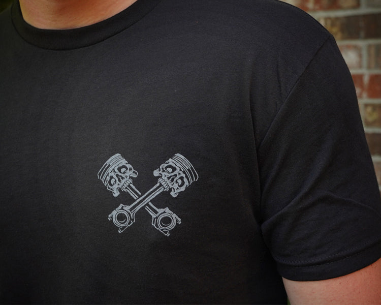 Black t-shirt with gray piston design, against a brick wall background.