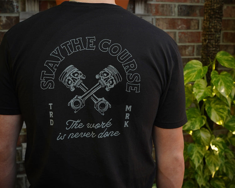 Person wearing a black t-shirt with a graphic design and text against a brick wall and plant background