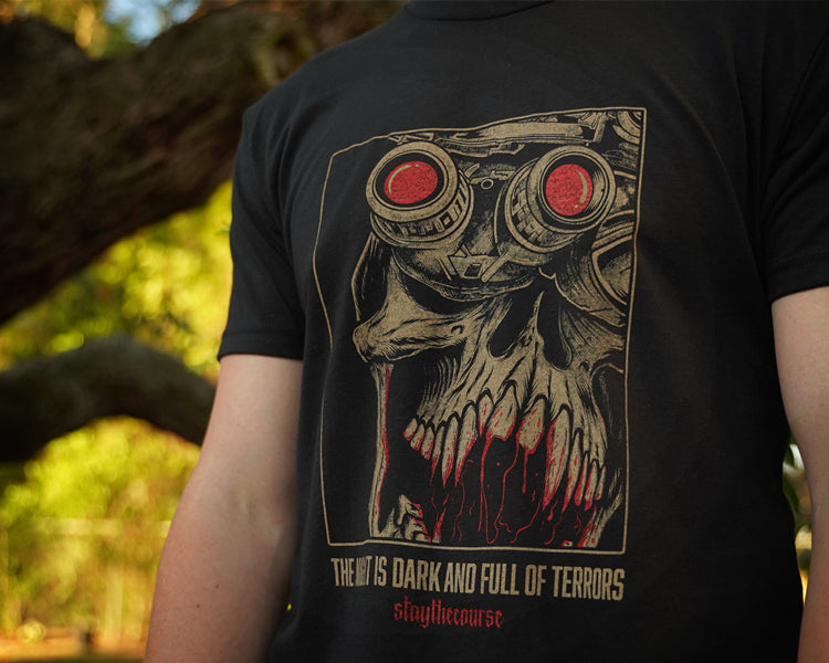 Person wearing a black t-shirt with tan and red graphic print and the words "the night is dark and full of terrors", standing in front of a tree