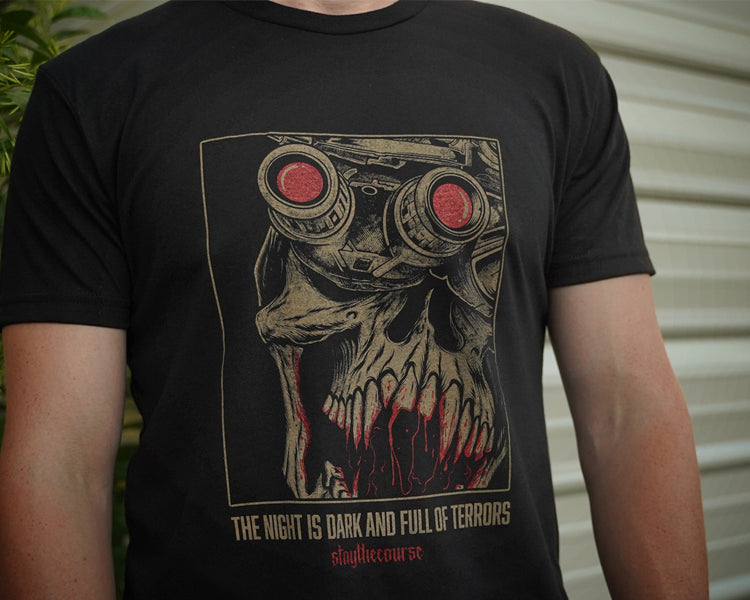 Person wearing a black t-shirt with tan and red graphic print and the words "the night is dark and full of terrors", standing in front of a building.