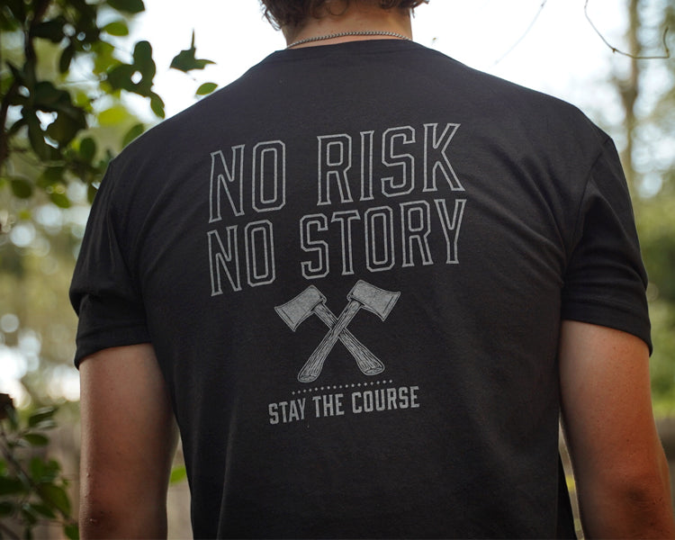 Person wearing a black t-shirt with 'No Risk No Story' text and graphic, outdoors.