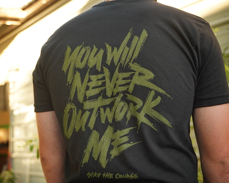 Person wearing a black t-shirt with green graphic print that says "you will never outwork me, stay the course", standing in front of a building.