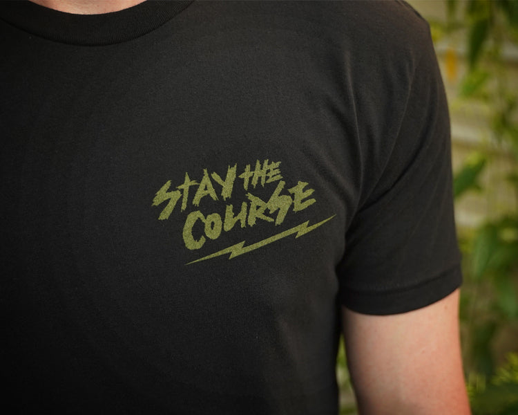 Person wearing a black t-shirt with green graphic print that says "stay the course", standing in front of a building.