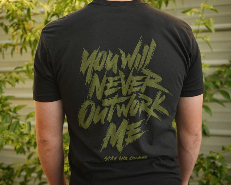 Person wearing a black t-shirt with green graphic print that says "you will never outwork me, stay the course", standing in front of a building.