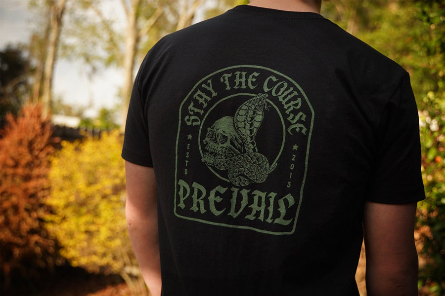 Person wearing a black t-shirt with a green "Stay the Course" "Prevail" graphic and text outdoors