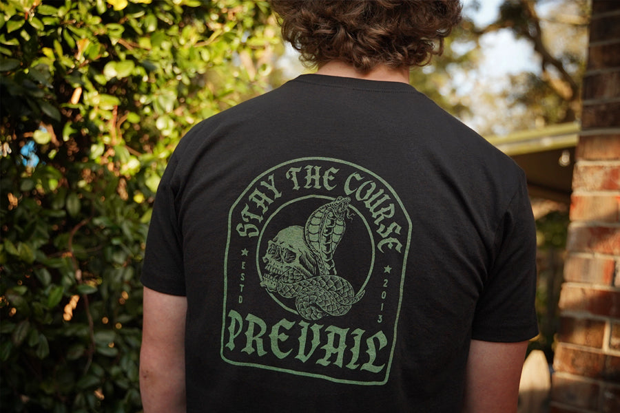 Person wearing a black t-shirt with hand printed green graphic "Stay the Course" and "Prevail" text outdoors