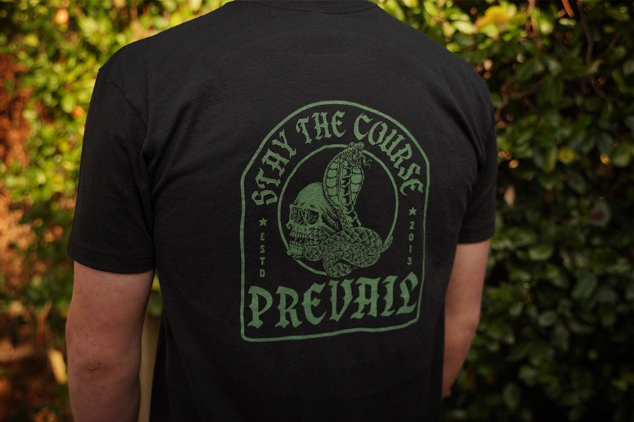 Person wearing a black t-shirt with green "Stay the Course" "Prevail" text and logo against a blurred green background