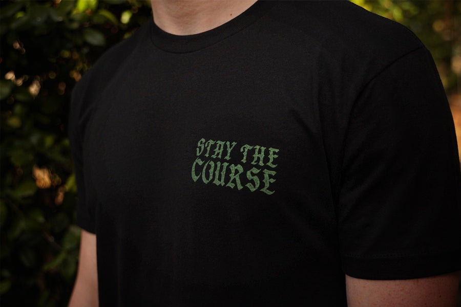 Black t-shirt with 'Stay the Course' text in green on a blurred natural background