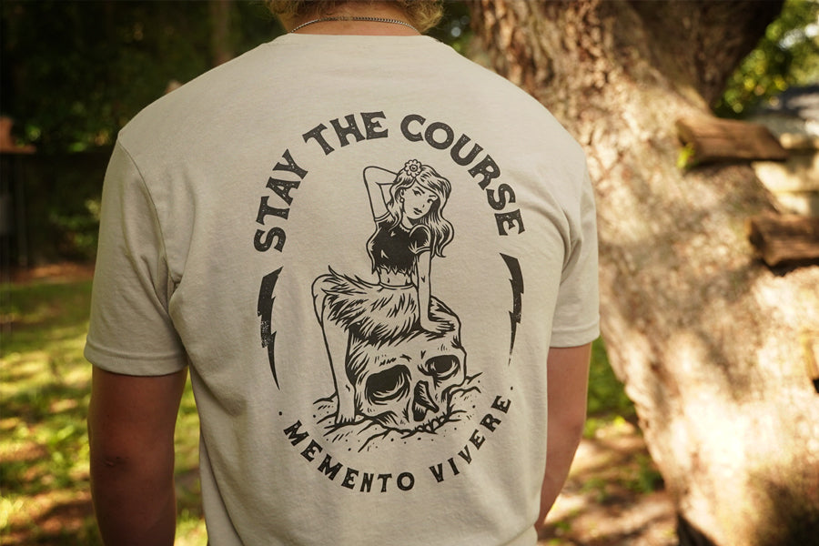 Person wearing a t-shirt with a graphic design and text 'Stay the Course Memento Vivere' outdoors.